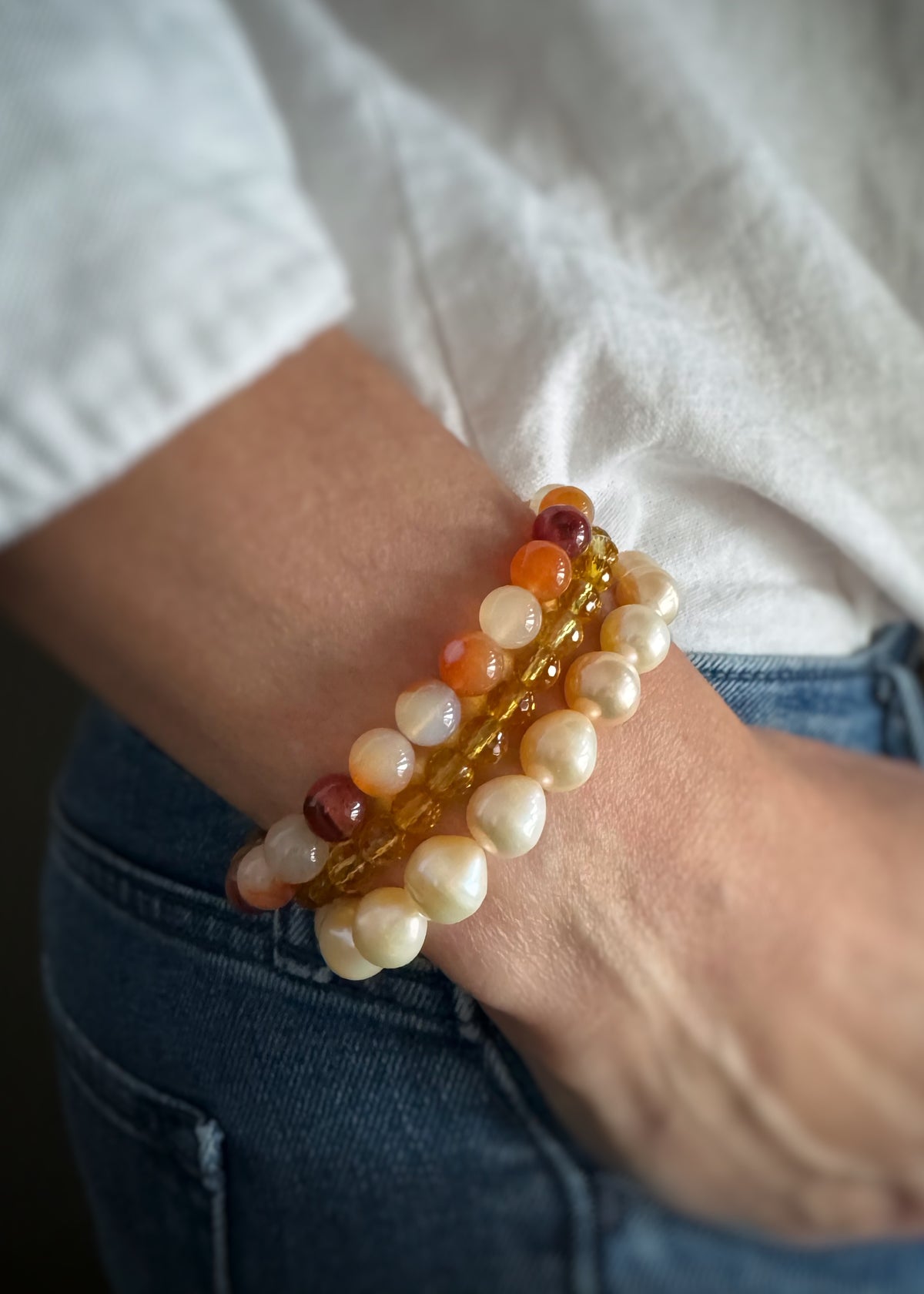 Close-up of a wrist wearing three bracelets; a carnelian stretch bracelet, a round faceted citrine bracelet, and a golden pearl stretch braceket against a white shirt and blue jeans background.