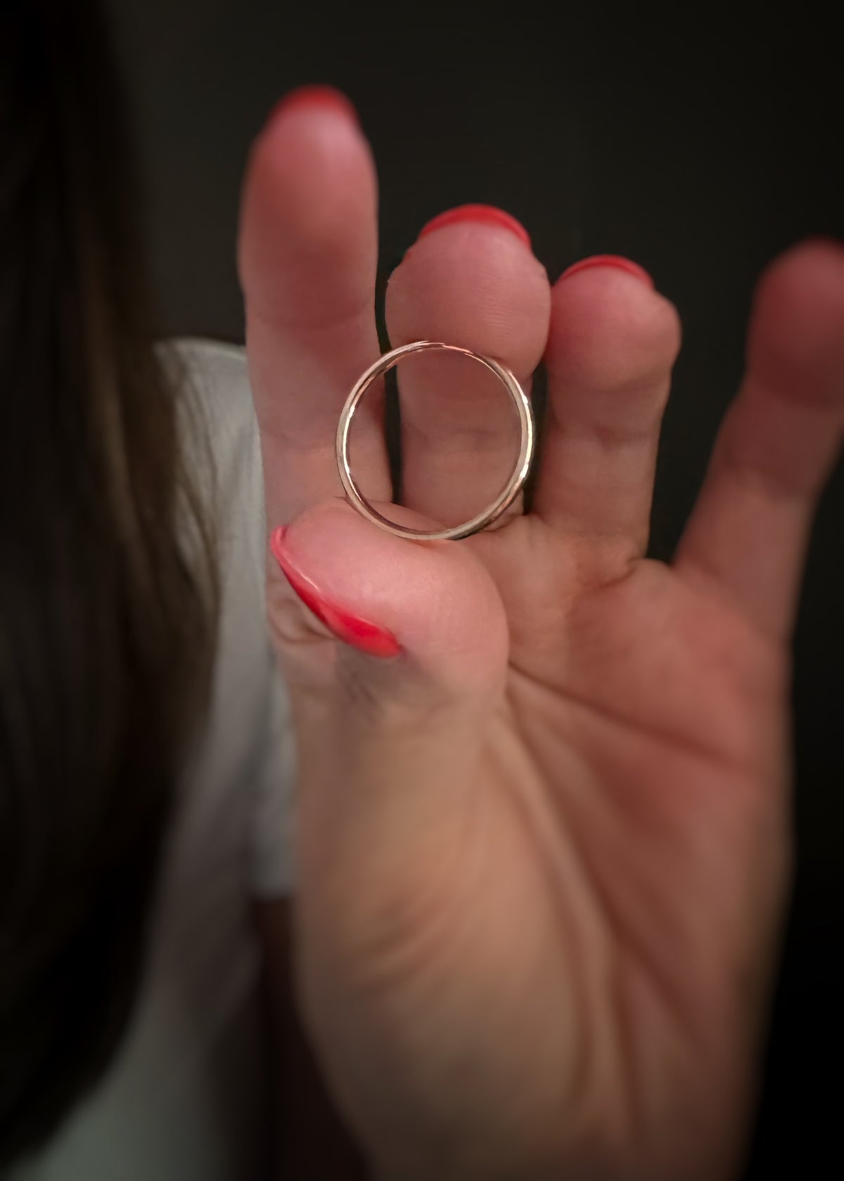 A close-up of a person's hand with red-painted fingernails holding a thin, polished 14k rose gold band ring against a dark background.