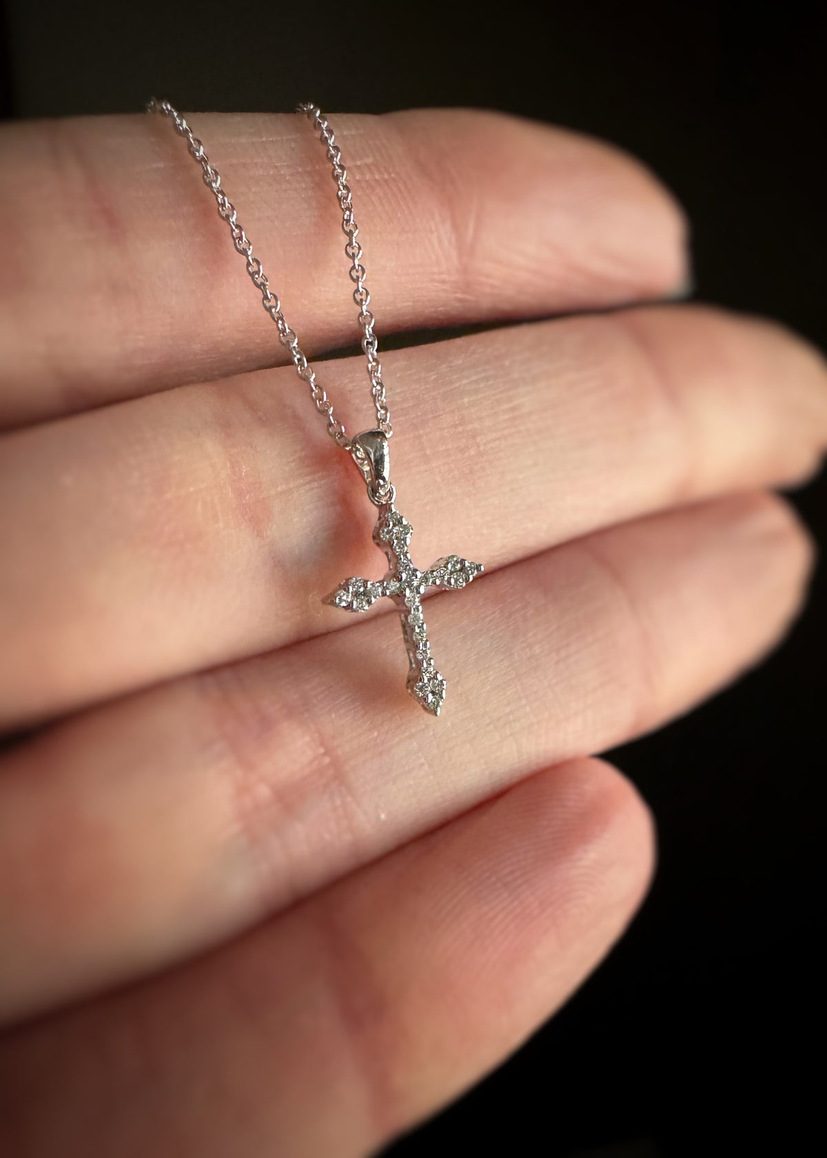 white gold cross necklace set with diamonds held between fingers against a dark background