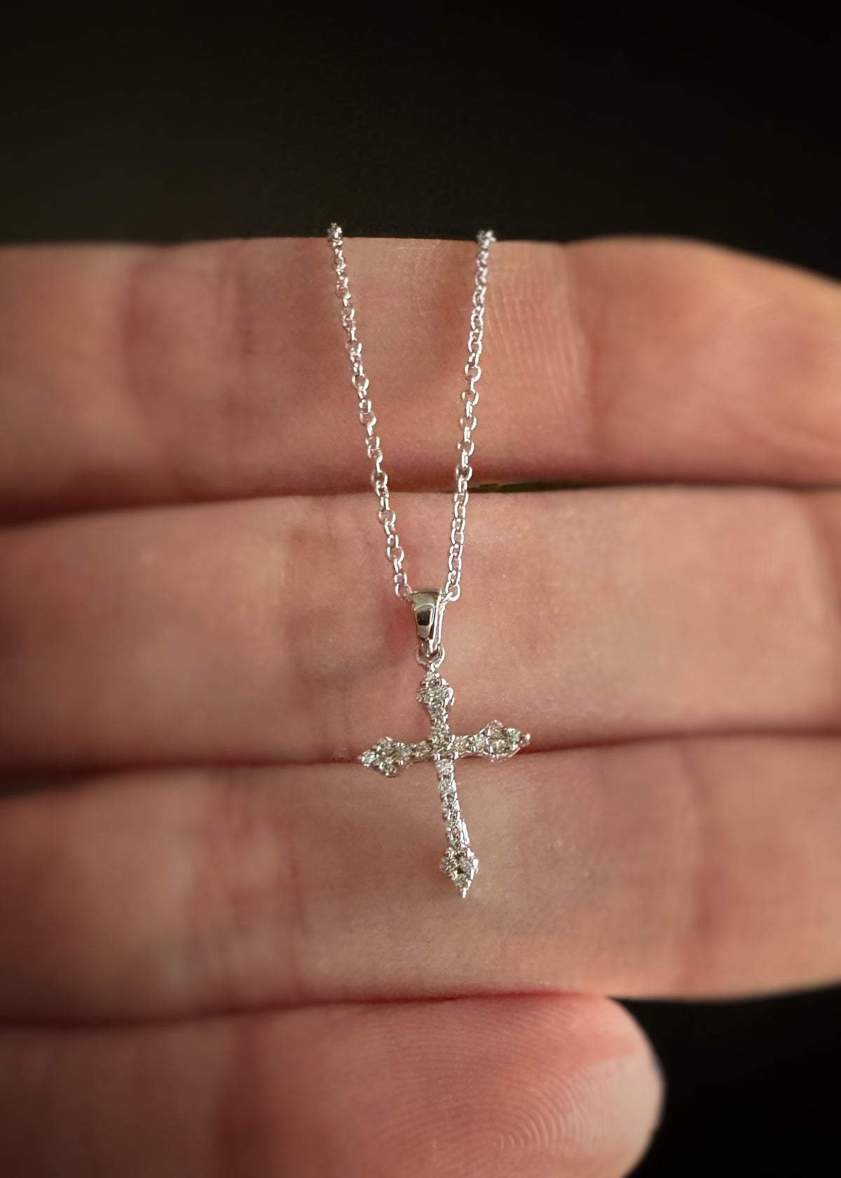 close up of a 14k white gold cross pendant set with round diamonds held between fingers against a dark background