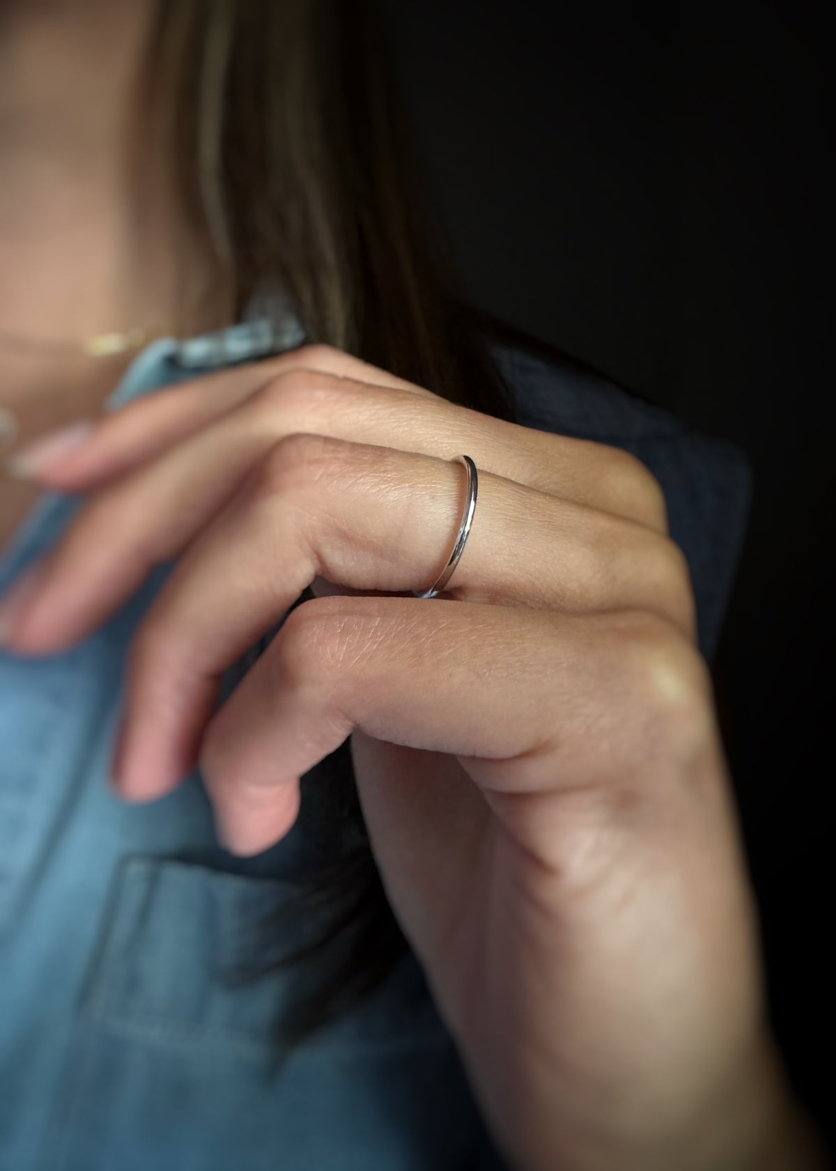 A close-up of a person's hand wearing a thin, polished 14k white gold band ring, dressed in a blue denim top against a dark background.