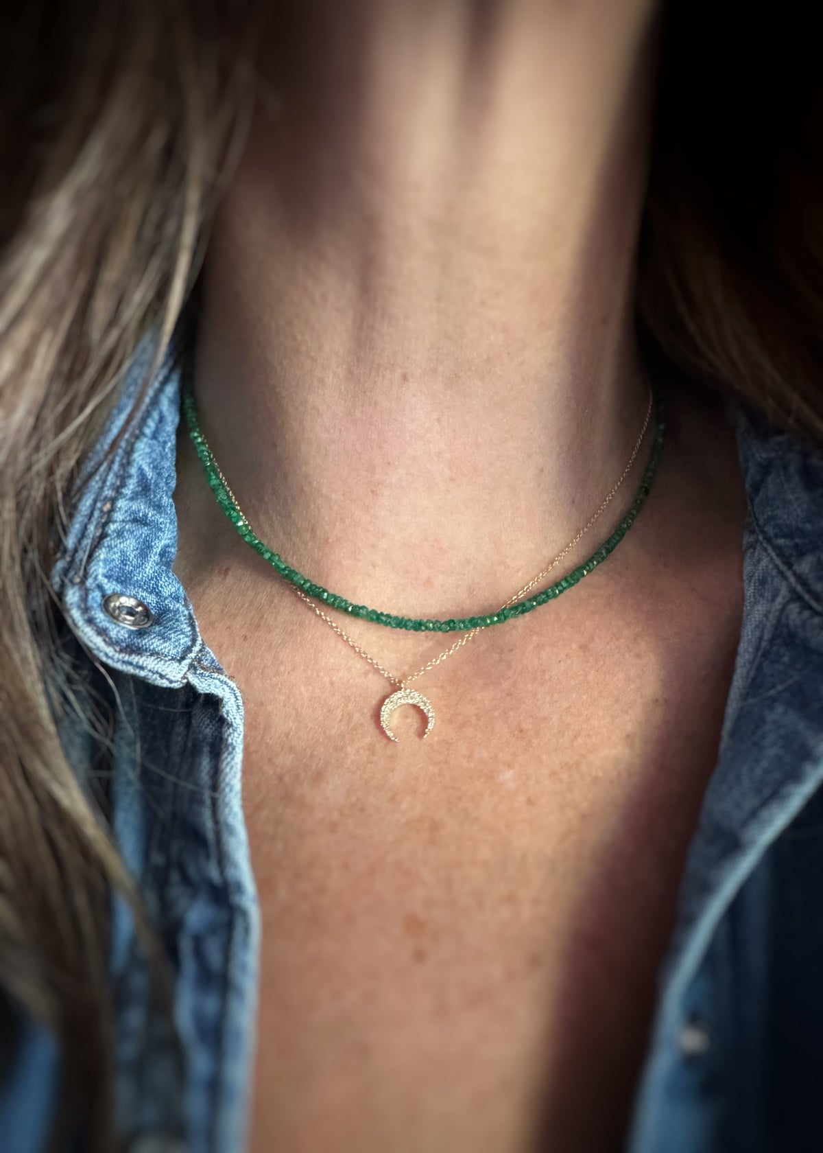 Close-up of a person's neck and upper chest wearing two layered necklaces: one green emerald bead and one delicate gold necklace with a diamond crescent moon pendant.