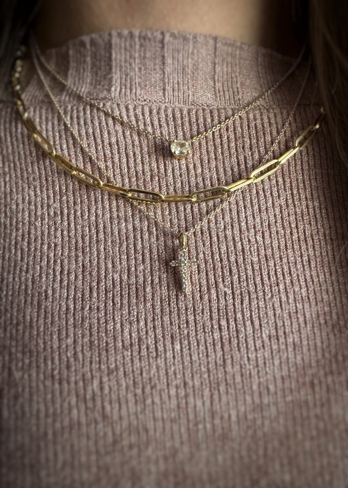 three gold necklaces stacked on a person's neck; one oval diamond necklace, a heavy paper-clip chain, and a yellow gold diamond cross charm on a textured brown surface