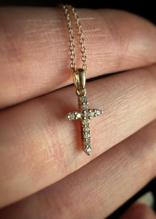 Yellow gold and diamond cross pendant on a chain held between fingers.