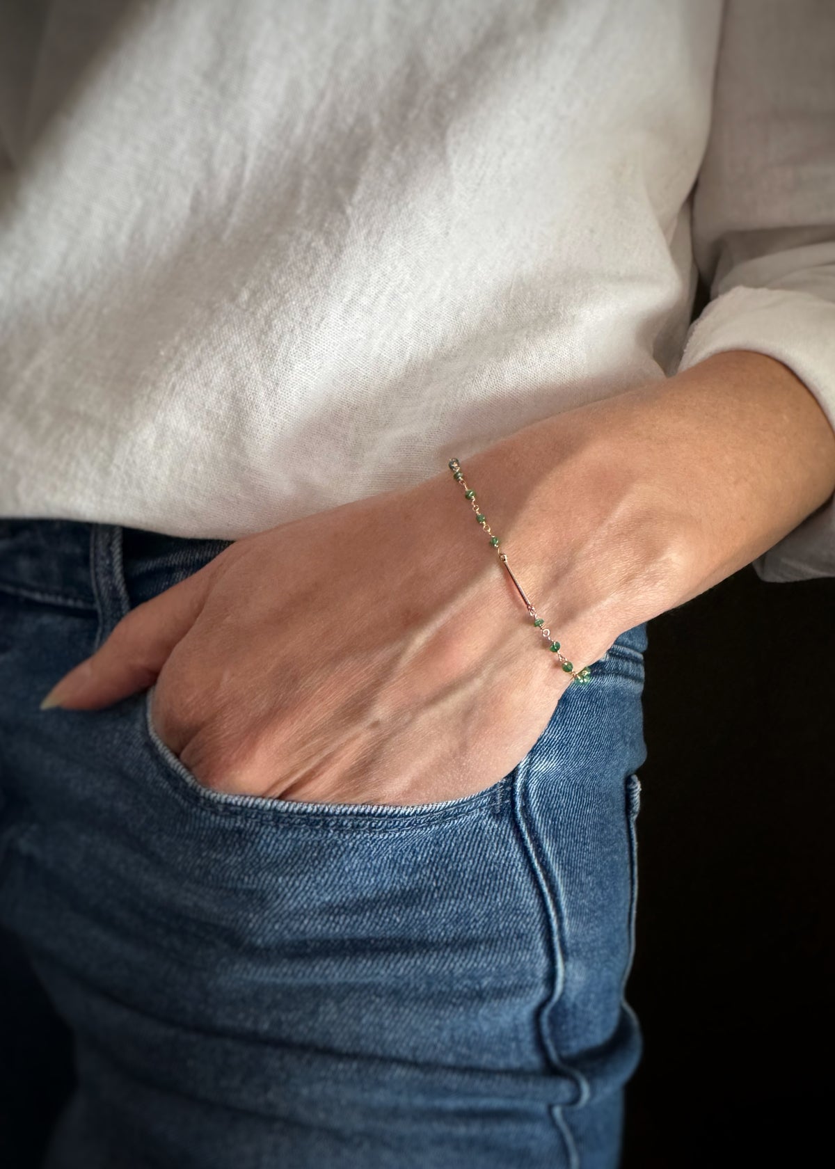 Person wearing a fine gold bracelet with emerald beads woven in the chain and a plain gold bar in the bracelet's center on a plain background