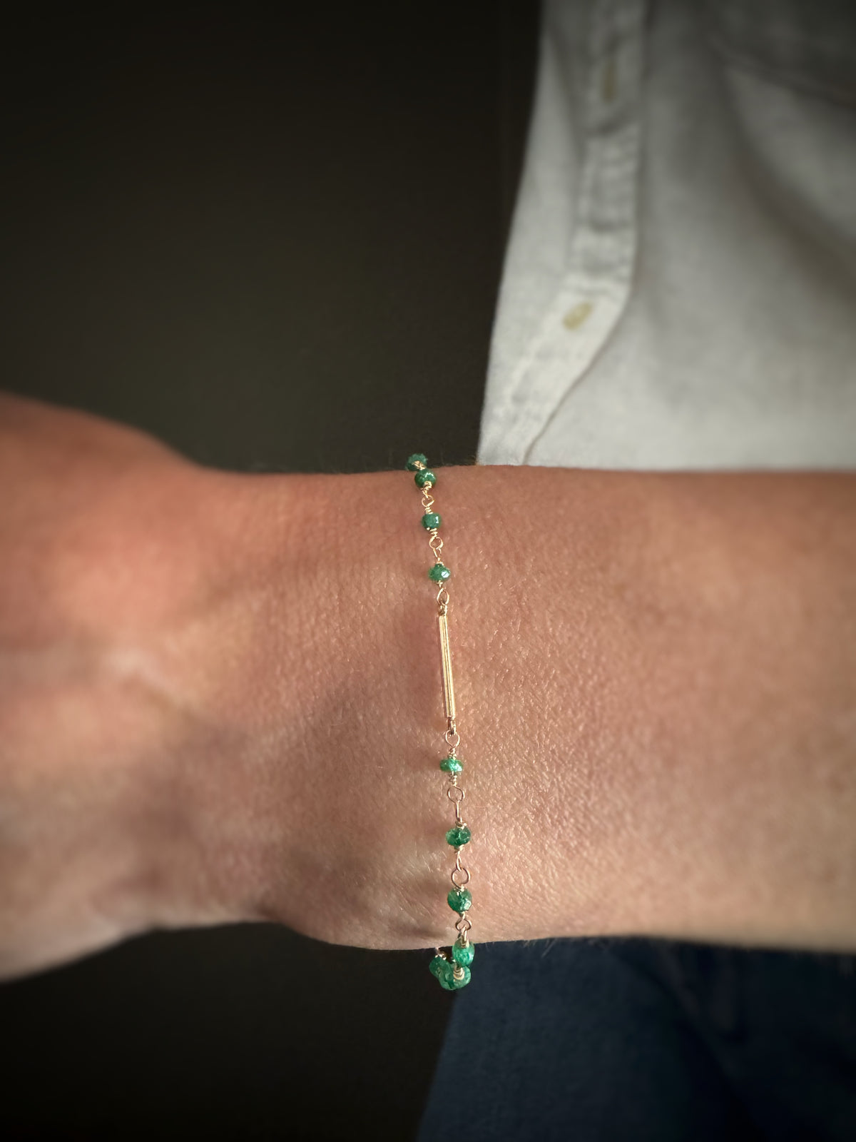 yellow gold bracelet with green emerald beads  and a gold bar in the center on a wrist against a neutral background
