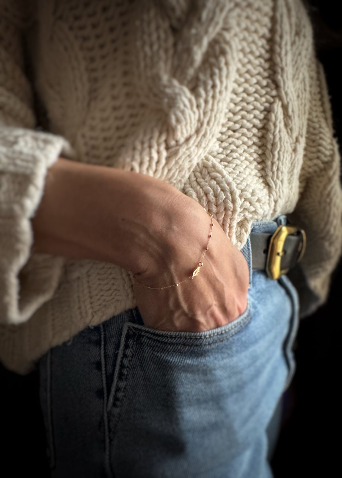Close-up of a yellow gold rosary chain bracelet on a person wearing a beige knitted sweater and blue jeans with a blurred background