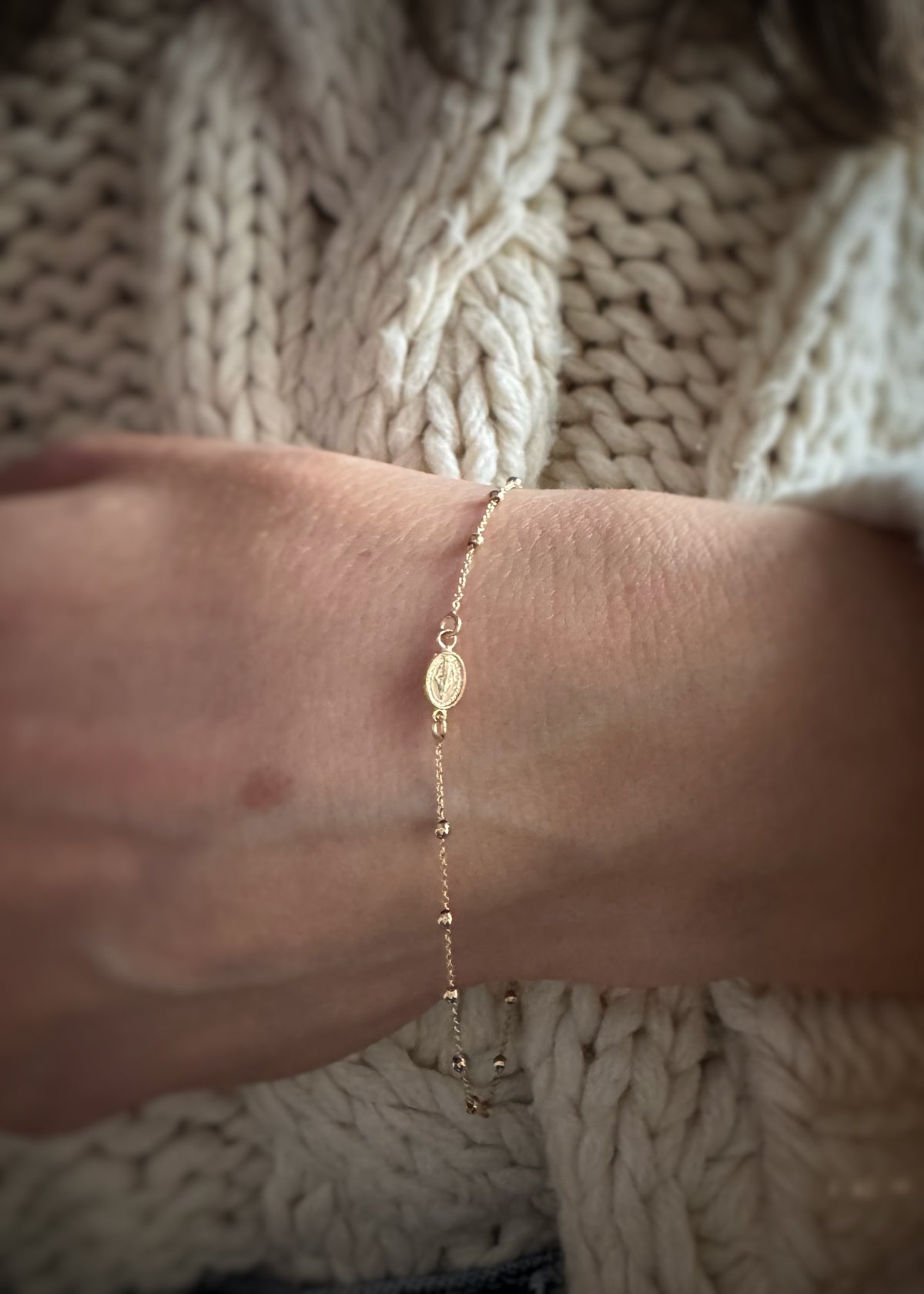 14k yellow gold rosary chain bracelet on a wrist with a textured beige sweater background