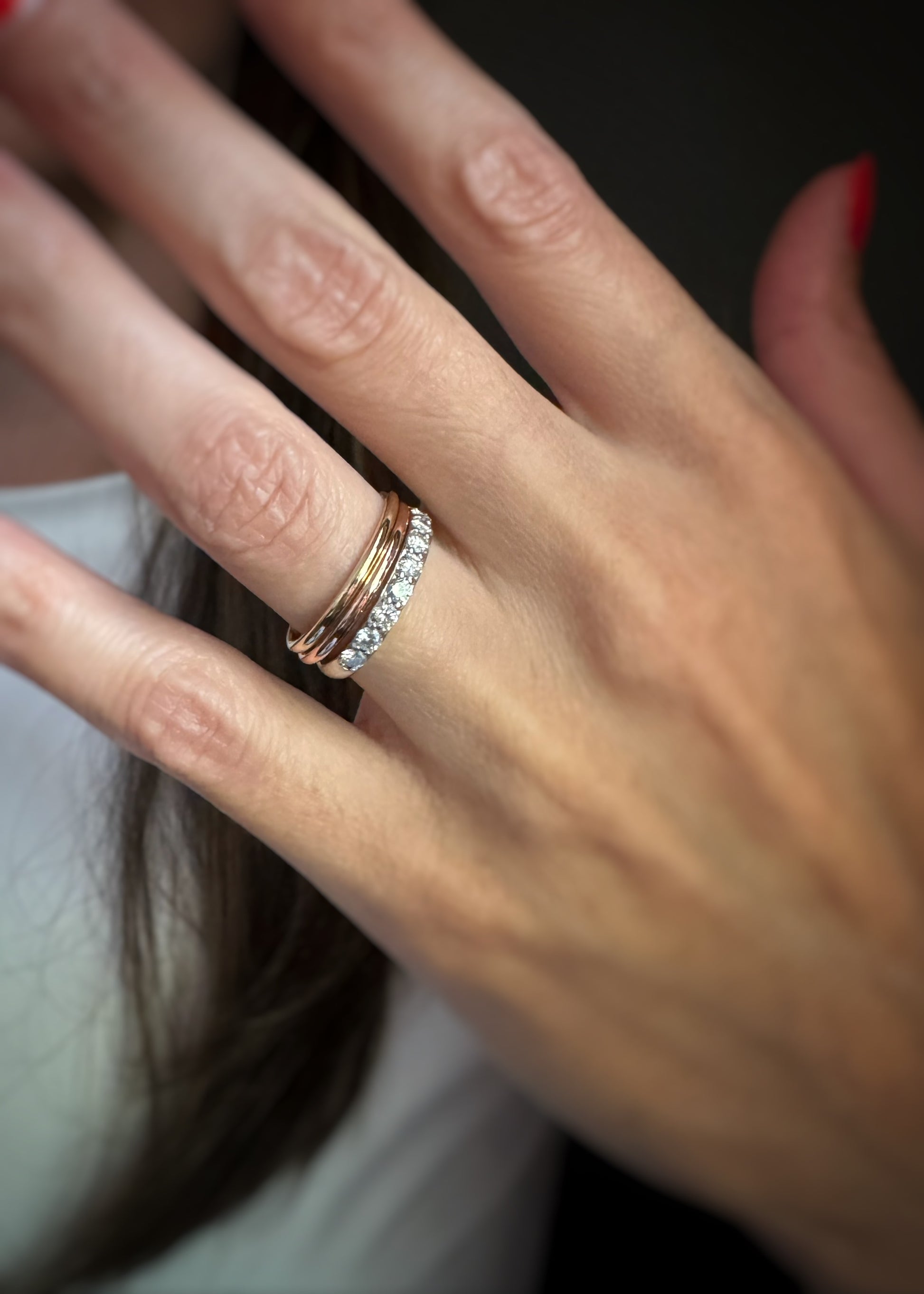 A close-up of a hand wearing two slim rings: one rose gold band and one white gold or platinum band set with small clear stones, against a dark background.