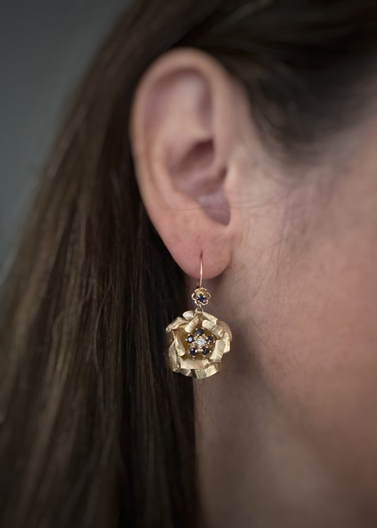 A close-up of a person's ear wearing a textured gold sapphire and diamond flower dangle earring.