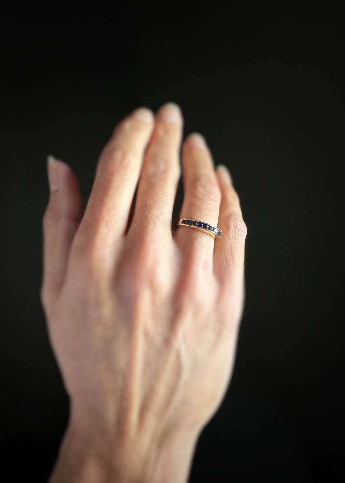 Hand wearing a wedding band with blue sapphires channel set across the band on a dark background