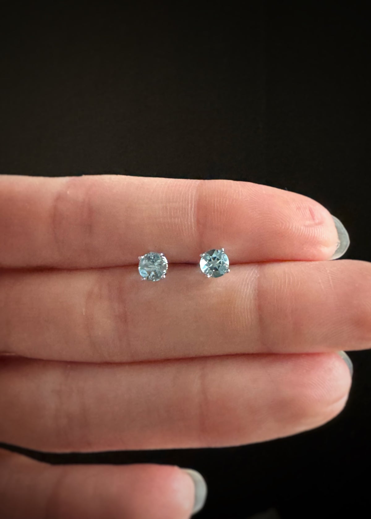 Two sky blue topaz gemstone stud earrings set in 14k gold held between fingers against a black background