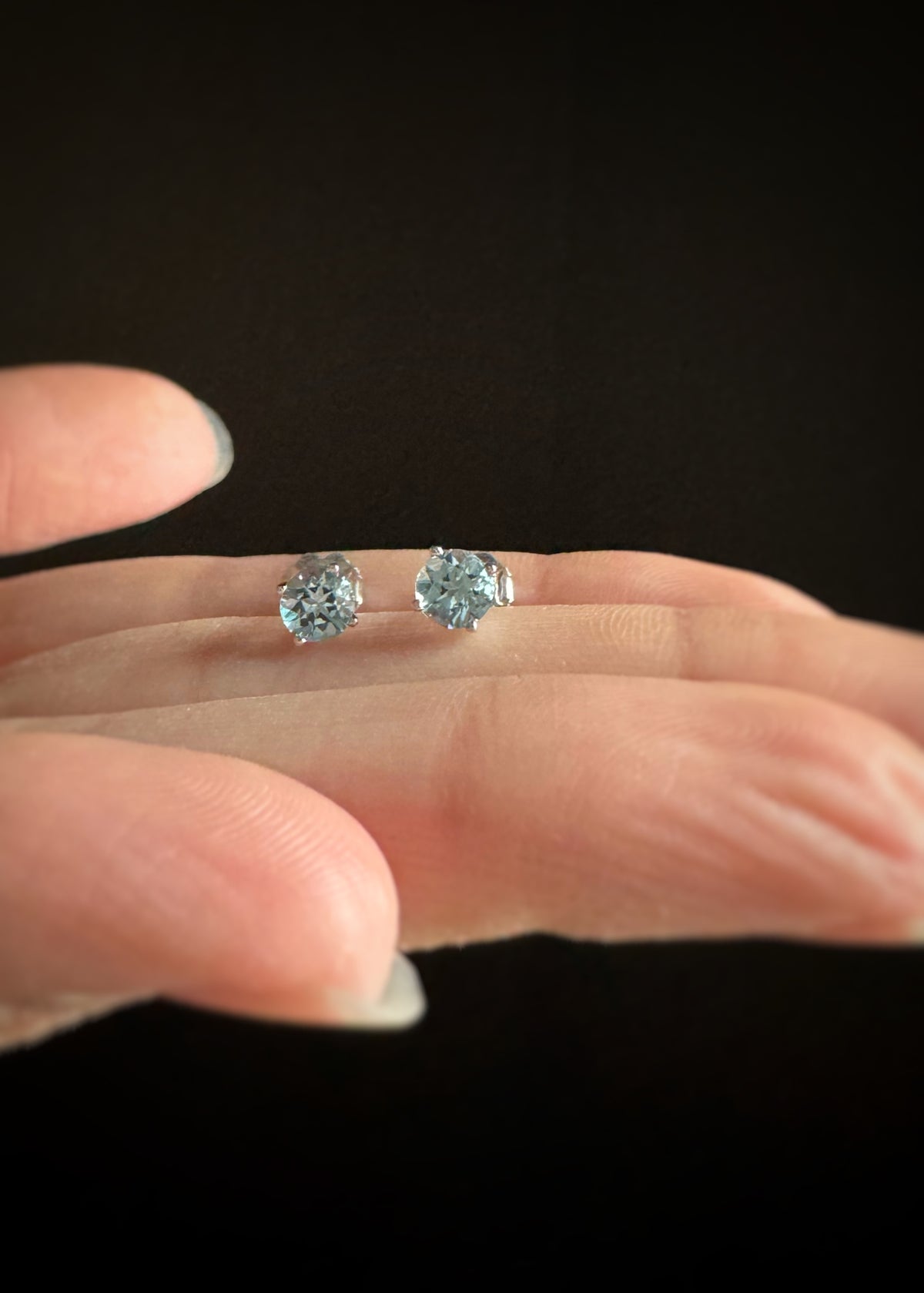 Two round cut blue topaz stud earrings set in 14k gold held between fingers against a dark background