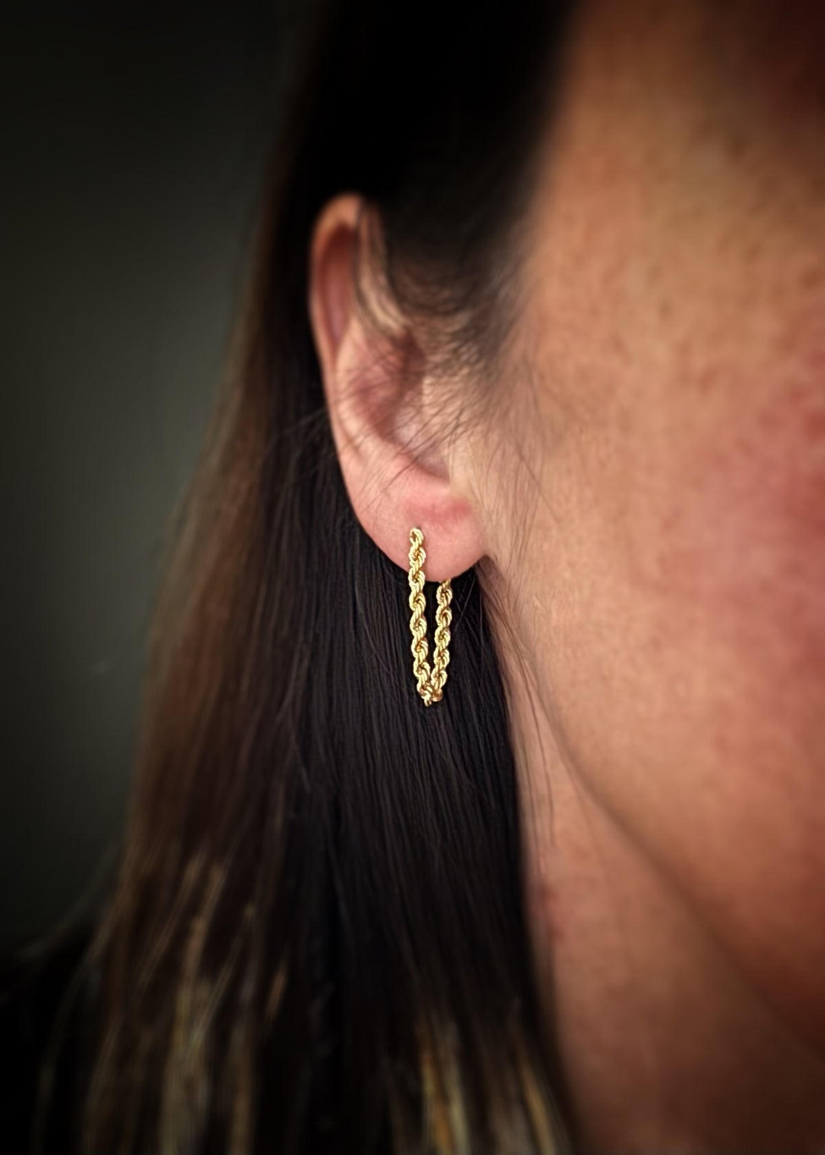 Close-up of a person wearing a 14 karat gold chain hoop earring with a dark background