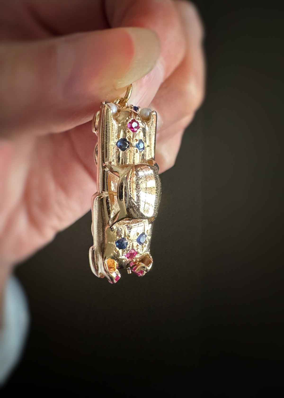 14 karat yellow gold classic car-shaped charm or pendant set with blue sapphires, red rubies, and white pearls with colorful gemstones held by a hand against a dark background