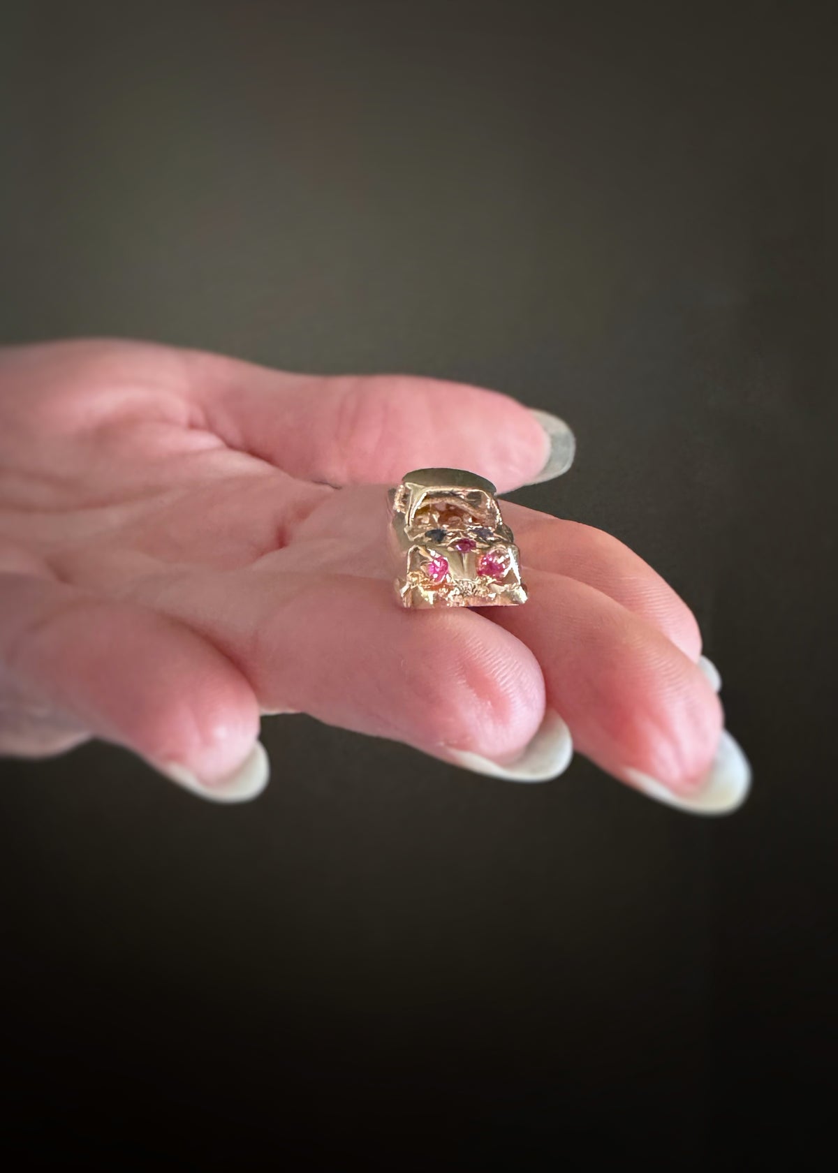 yellow gold vintage car charm showing the trunk of the car set with red rubies as taillights held on a finger against a dark background