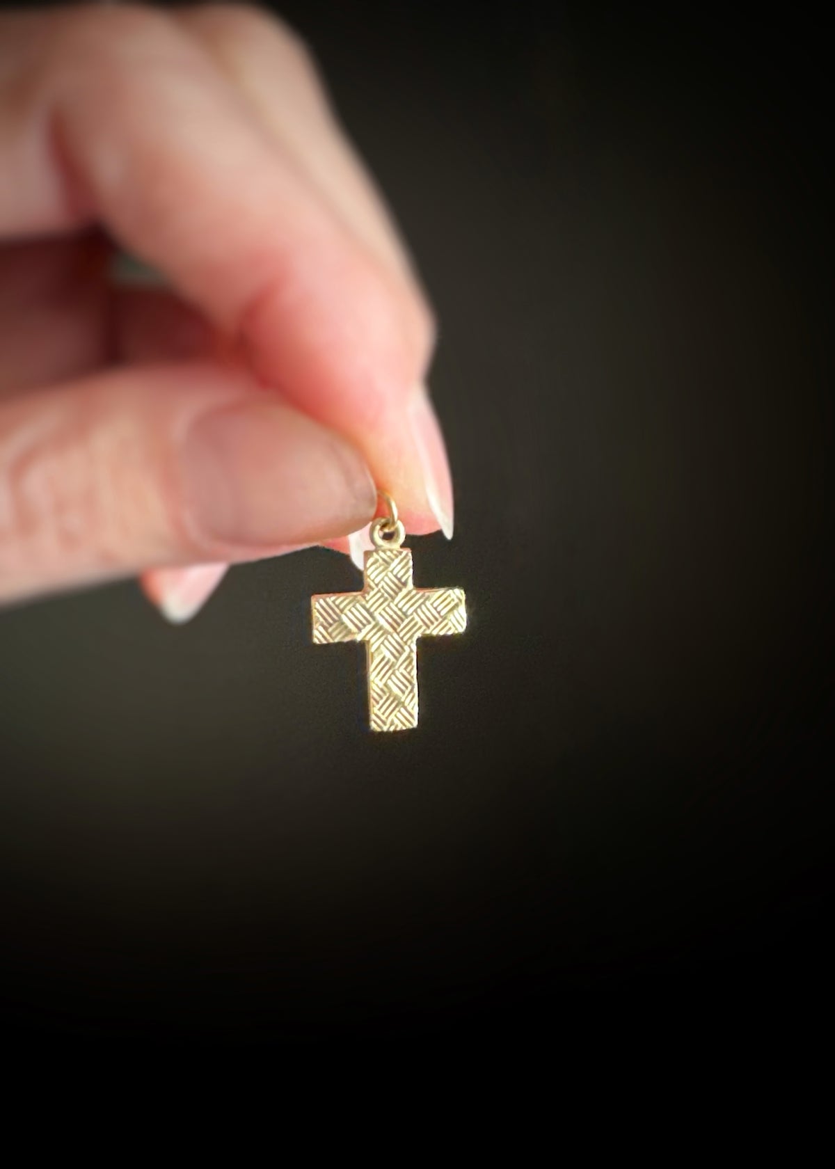 14k yellow gold cross pendant with thatch like texture held by a hand against a dark background