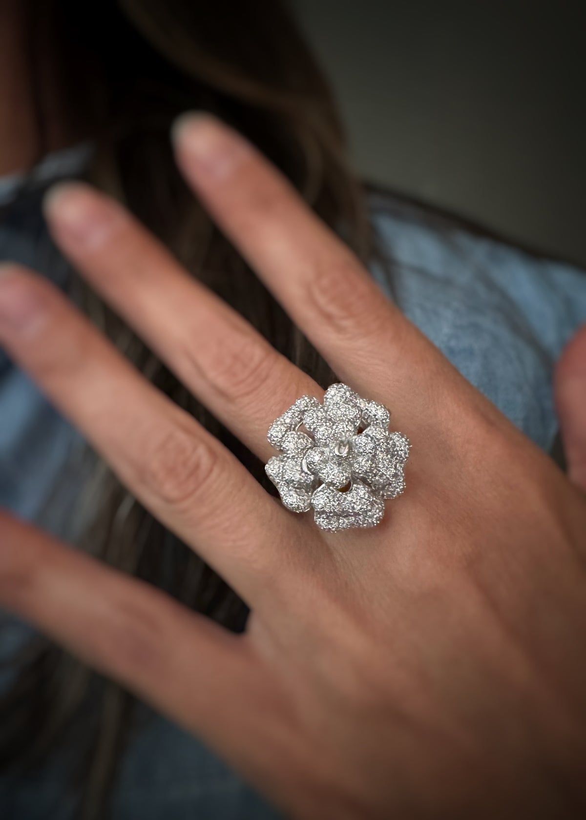 A close-up of a hand wearing a large, detailed floral cocktail ring with clusters of small diamonds set on each 18kt white gold petal, with the shoulder of the model in the soft-focus background.