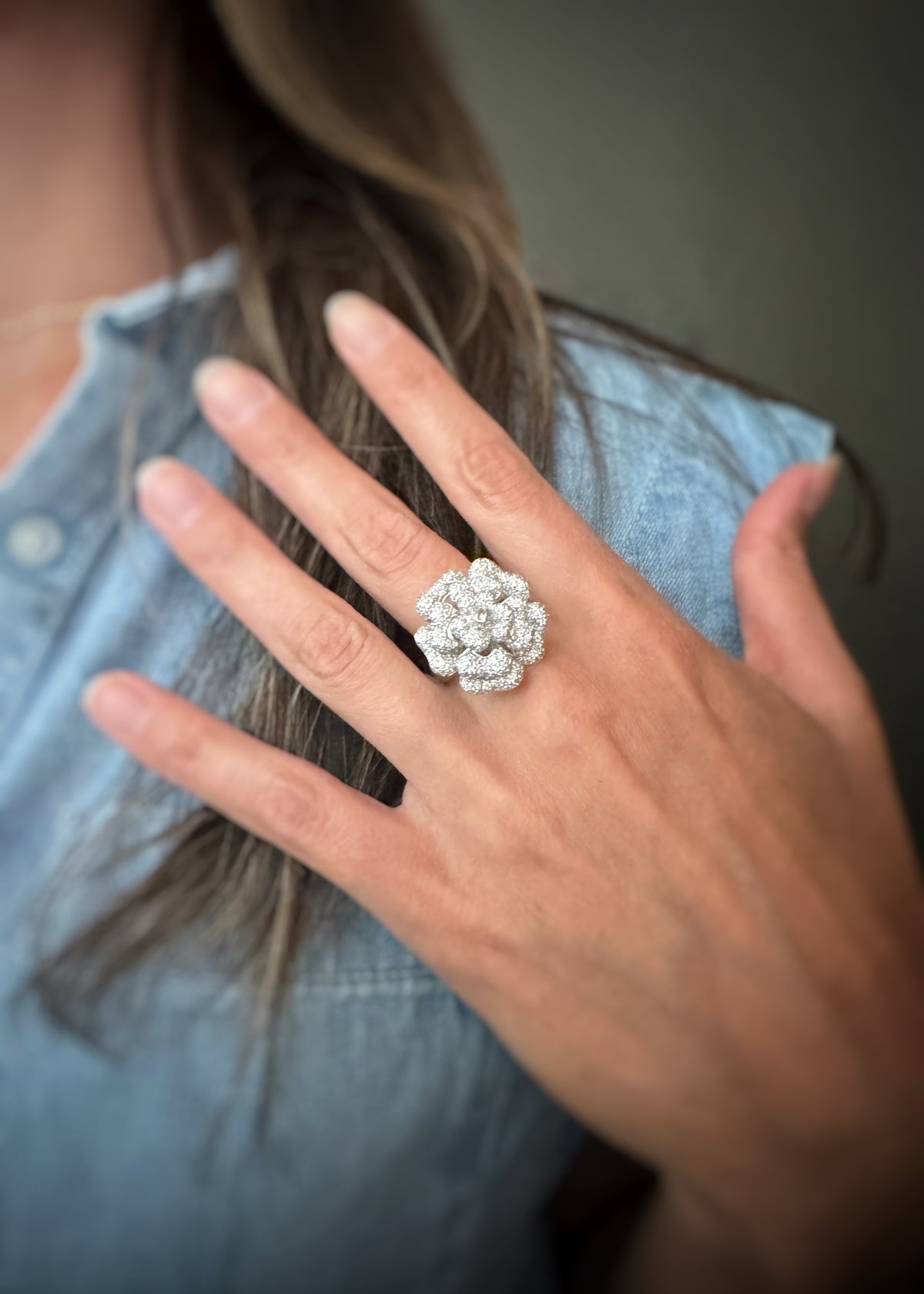 A close-up of a person's hand wearing a large, intricate 18k white gold ring with a floral, cluster-like design, set against a soft-focus background of the model's hair and a denim shirt.