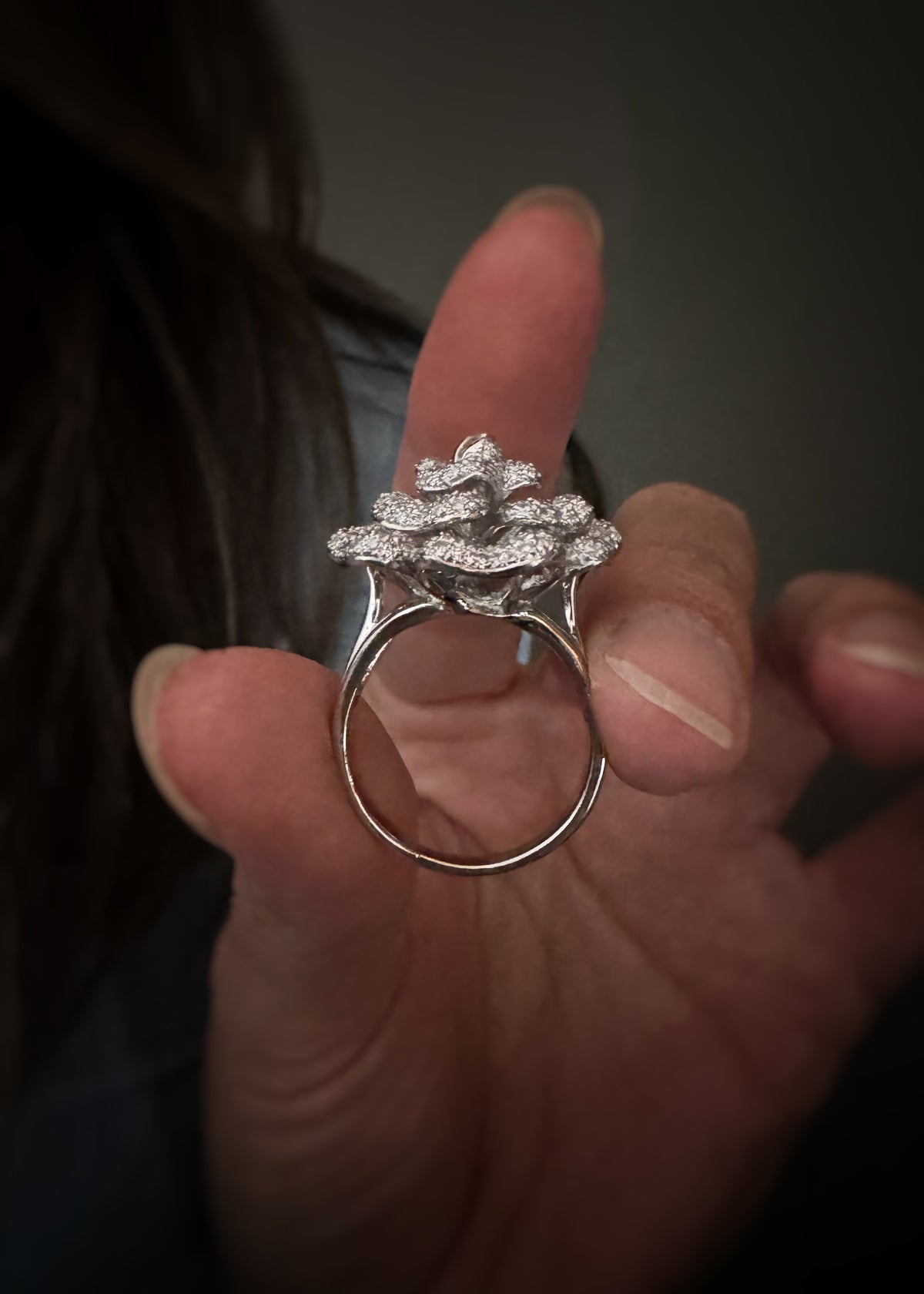 A profile view of an 18k white gold ring with small round diamonds pave set on each petal. The ring is held between fingers against a dark, blurred background.
