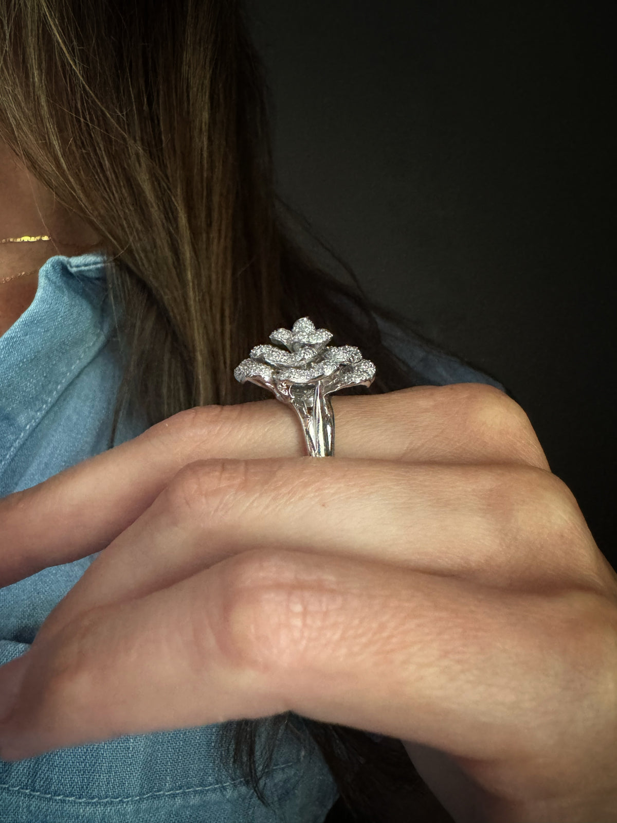 The side view of a tall 18k white gold ring with a sculptural, textured floral top set with small natural diamonds, worn on a person's finger. The person is wearing a light blue denim top against a dark background.
