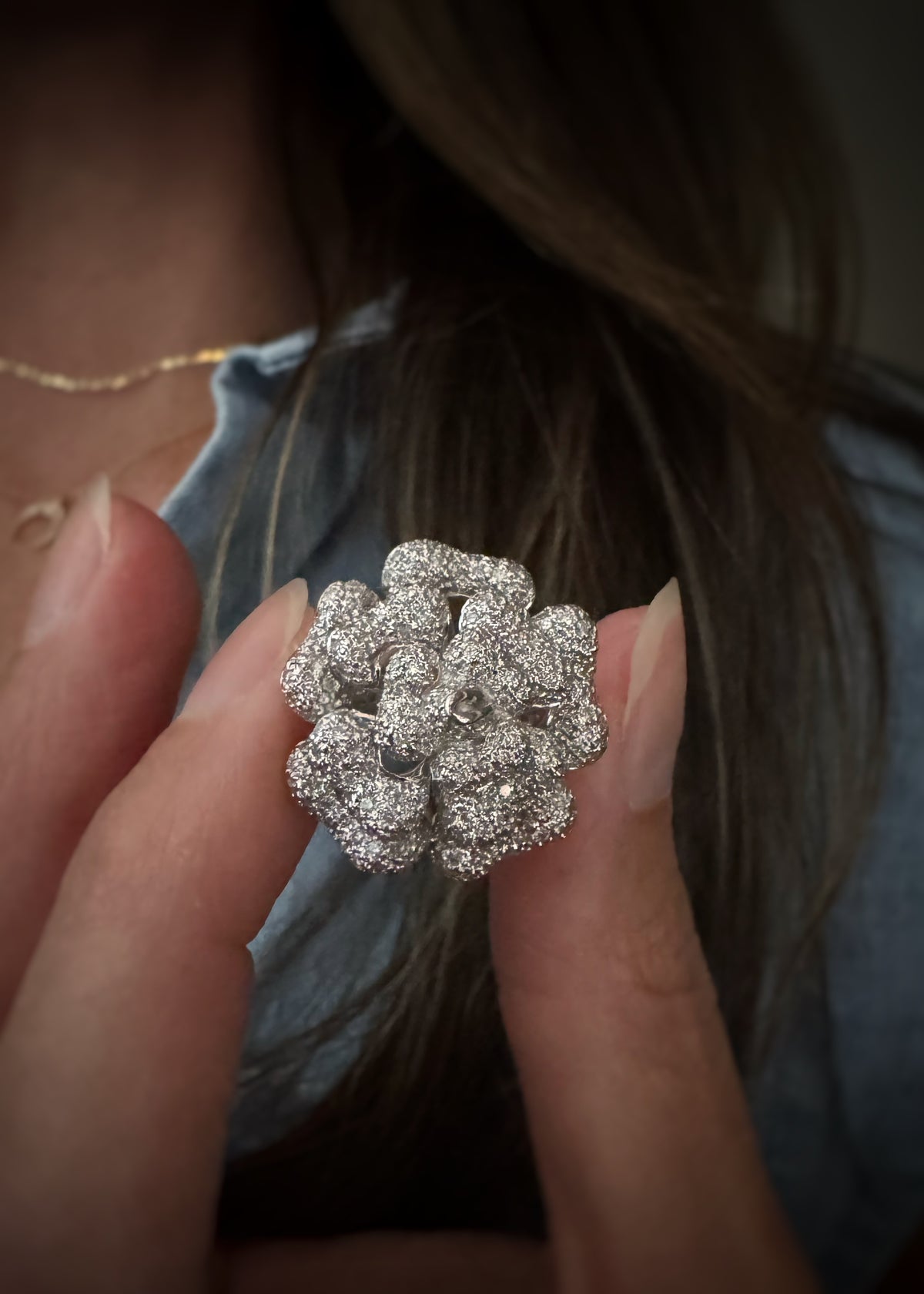 A close-up of a person holding an 18k white gold flower-shaped cocktail ring with pavé-set natural diamonds. The soft blurred background shows dark hair and a blue denim top.