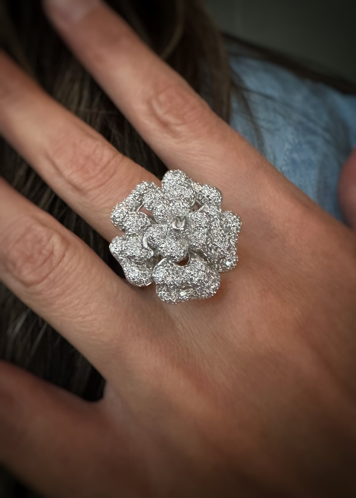 A close-up of a hand wearing a large, detailed floral cocktail ring with clusters of small diamonds set on each 18kt white gold petal, with the shoulder of the model in the soft-focus background.