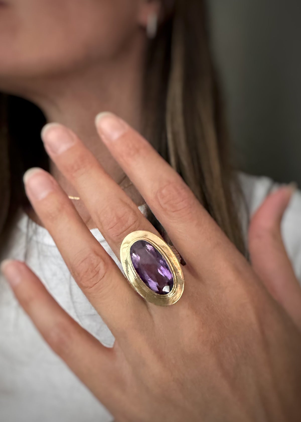 A close-up of a woman's hand wearing a gold ring with a large oval purple amethyst gemstone, set against a soft-focus background of her face and white shirt.