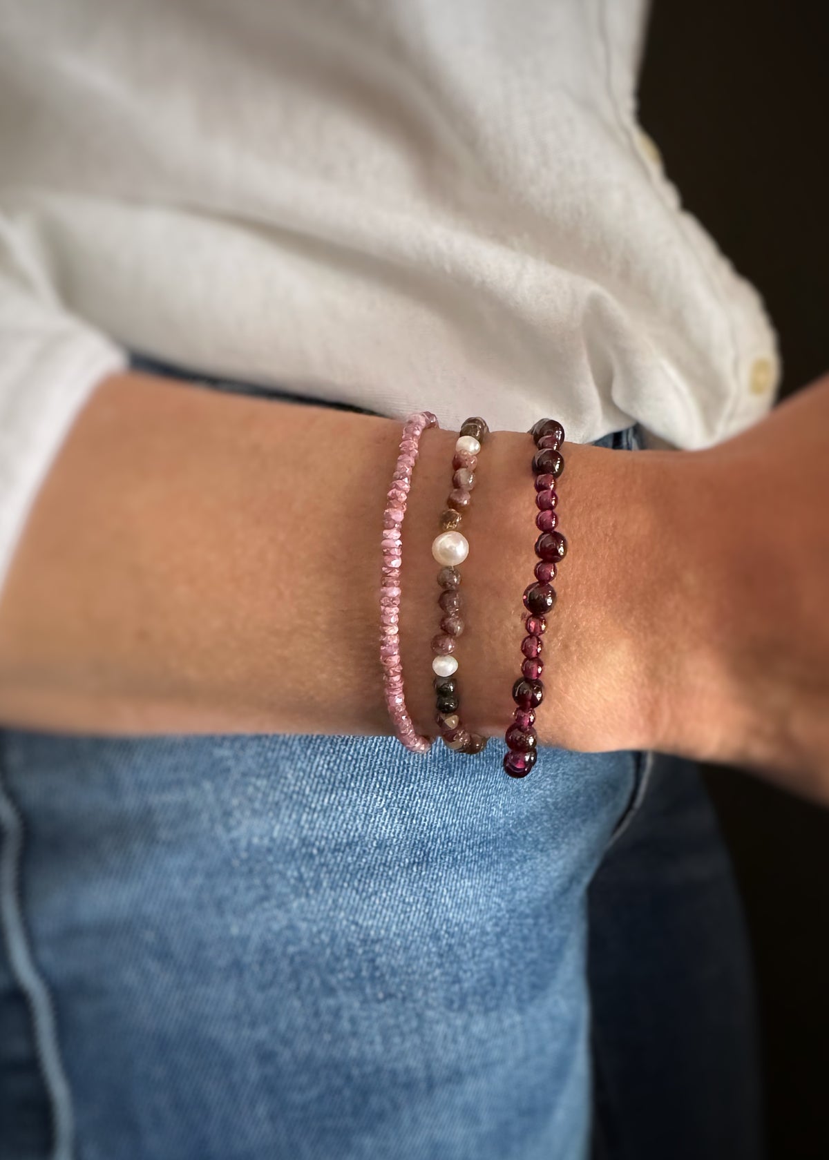 Close-up of a wrist wearing three beaded bracelets; a rough ruby elastic strand bracelet, tourmaline and pearl elastic strand bracelet, a smooth round garnet bead bracelet with a neutral background