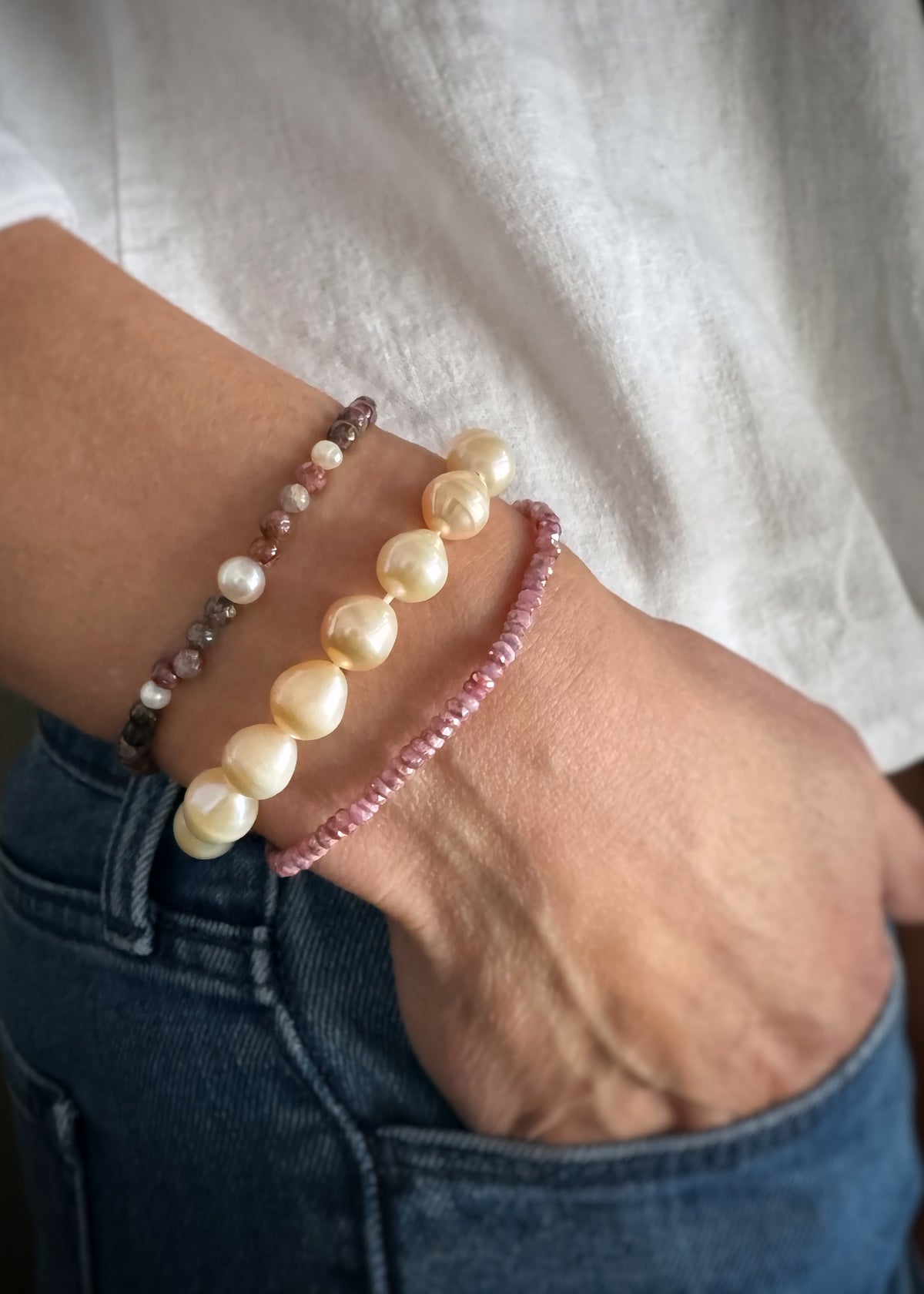 close up of a wrist with three elastic stretch bracelets; a mixed pink and green tourmaline bead bracelet, a golden baroque pearl bracelet, and light pink ruby rough bead bracelet.